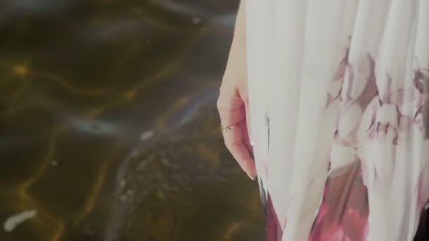 Pernas de uma jovem mulher de vestido branco com estampa floral andando ao longo do litoral — Vídeo de Stock