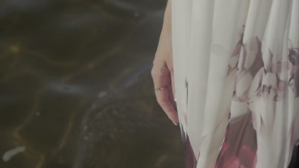 Ноги молодої жінки в білій сукні з квітковим принтом, що йде уздовж морського плаского кольору — стокове відео