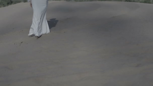 Πόδια του γυναίκα που περπατά στην παραλία αμμόλοφους σε λευκό φόρεμα, ενώ ήλιο μειώνεται σκιωδών αργή κίνηση εμπρός δείτε επίπεδη χρώμα — Αρχείο Βίντεο