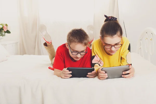 Dos niños juegan en tabletas tumbados en la cama — Foto de Stock