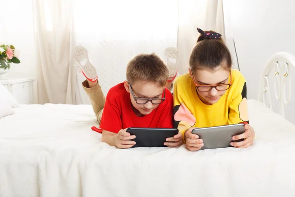 Двое детей играют на планшетах, лежащих на кровати — стоковое фото