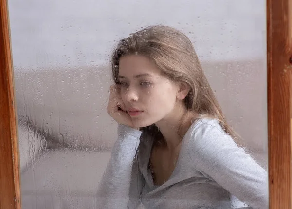 Piękna młoda kobieta smutna z powodu deszczowej pogody, wyglądająca przez okno i myśląca — Zdjęcie stockowe
