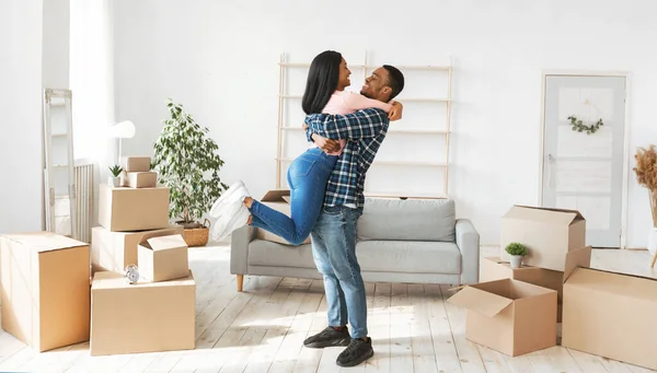 Conceito de mudança de casa. Feliz cara negro abraçando e levantando sua linda esposa em seu novo apartamento, panorama — Fotografia de Stock