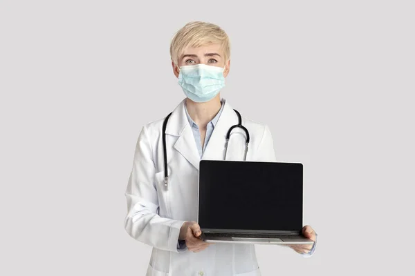 Vážná dospělá žena v bílém kabátě se stetoskopem a ochrannou maskou ukazuje notebook s prázdnou obrazovkou — Stock fotografie