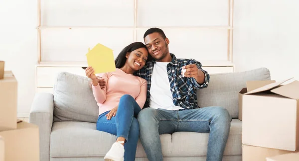День переїзду. Молода афроамериканська пара з паперовим будинком і ключем до свого нового дому сидить на дивані і посміхається — стокове фото