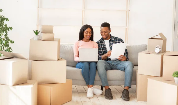 Νεαρό ζευγάρι Αφροαμερικανών κάθεται στον καναπέ ανάμεσα σε χαρτοκιβώτια με φορητό υπολογιστή και τη δημιουργία σχεδίου για το νέο σπίτι — Φωτογραφία Αρχείου