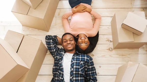 Wanita muda kulit hitam dengan suaminya yang tampan berbaring di antara kotak kardus di lantai rumah baru mereka, pemandangan atas — Stok Foto