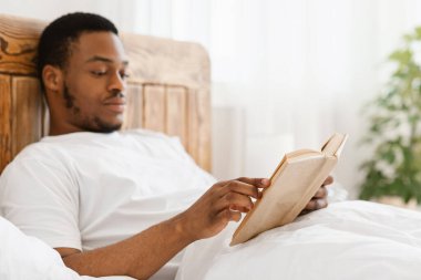 Afro-Amerikalı adam kitap okuyor. Sabahın zevkini çıkarıyor. Yatakta oturuyor.