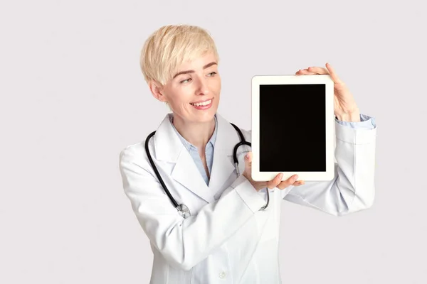 Szczęśliwy w średnim wieku dorosła kobieta lekarz w białym płaszczu ze stetoskopem pokazuje cyfrową tabletkę z pustym ekranem — Zdjęcie stockowe