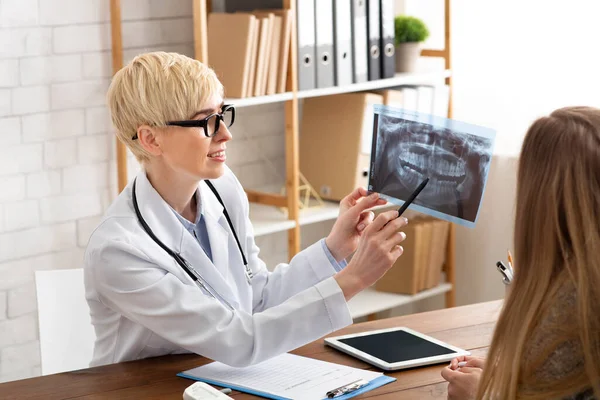 Beyaz önlüklü ve gözlüklü yetişkin kadın doktor hasta çene, diş ve teşhislerinin röntgenini gösteriyor. — Stok fotoğraf