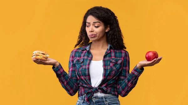 Hambre mujer negra sosteniendo hamburguesa y fruta de manzana — Foto de Stock