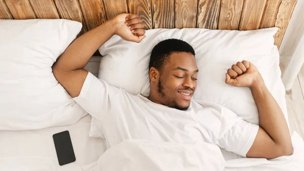 Κοιμημένος μαύρος άντρας ξυπνά στο κρεβάτι τεντώνοντας τα χέρια στην κρεβατοκάμαρα — Φωτογραφία Αρχείου