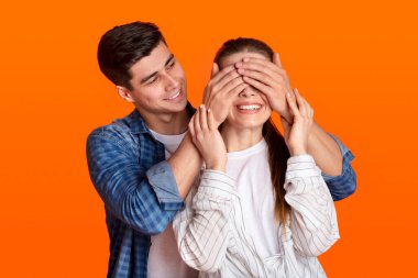 Young guy stands behind lady and closes eyes of smiling woman isolated on orange background clipart