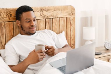 Siyah adam dizüstü bilgisayar kullanıyor. İnternette film izliyor. Yatakta yatıyor.