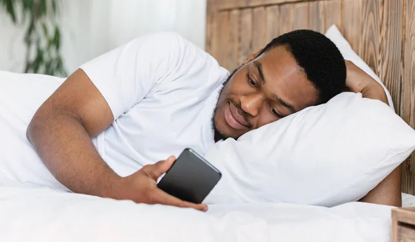 Africano americano cara usando celular deitado na cama no quarto — Fotografia de Stock