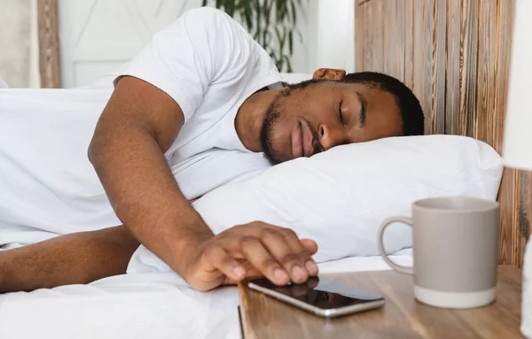 Hombre africano durmiendo a través del despertador en el teléfono inteligente acostado en la cama — Foto de Stock