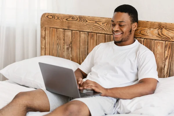 Μαύρος τύπος που εργάζεται σε φορητό υπολογιστή μακρινά σε απευθείας σύνδεση κάθεται στην κρεβατοκάμαρα — Φωτογραφία Αρχείου