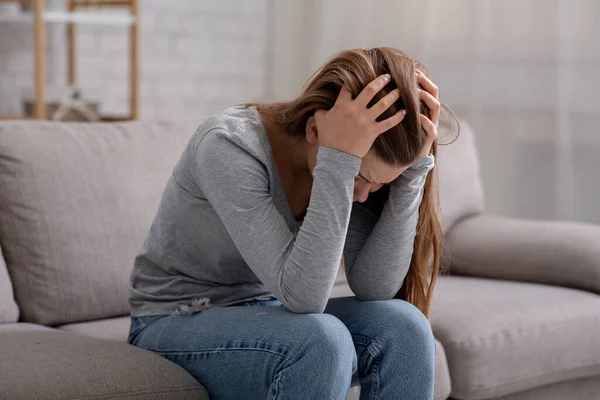 Нещасна молода жінка з головою в руках сидить на дивані, плаче і відчуває себе пригніченою — стокове фото