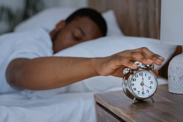 El hombre africano despierta apagando el reloj despertador acostado en la cama —  Fotos de Stock