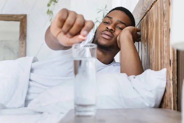 Zieke Afrikaanse man zetten pil in glas liggend in bed — Stockfoto