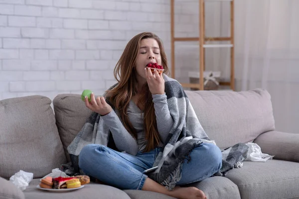 Sad young lady coping with depression or stress by eating sweets on sofa at home