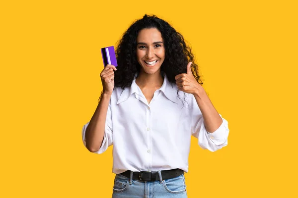 Positivo morena mulher segurando cartão de crédito e gestos polegar para cima no câmara — Fotografia de Stock