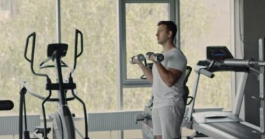 Spor salonunda, iç mekanda, yan görüş, spor yaşam tarzı konseptinde dambıllarla egzersiz yapan motive olmuş güçlü bir adam.