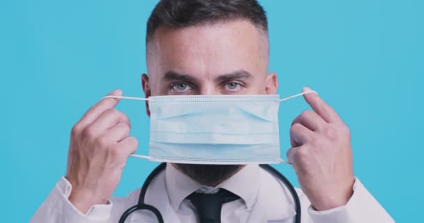 Nuevo concepto normal. Médico maduro poniéndose máscara médica protectora, retrato de estudio sobre fondo azul — Vídeos de Stock