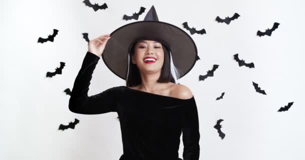Портрет милої азіатки, яка позує в костюмі відьми Хелловін і сміється над білою стіною з прикрасою кажанів — стокове відео