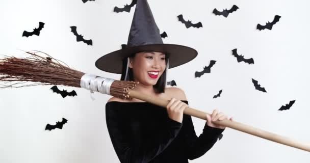 Joven asiática mujer en halloween encanto traje posando con escoba, blanco estudio fondo con murciélagos decoraciones — Vídeo de stock