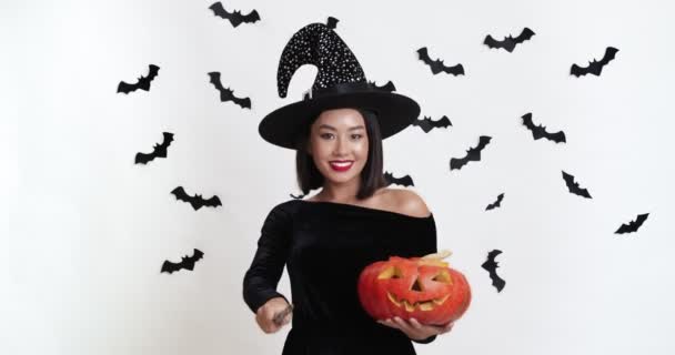Geleneksel Cadılar Bayramı. Cadı kostümlü neşeli Asyalı kadın elinde balkabağı oyması ve sihirli değnekle el kol hareketi yapıyor. — Stok video