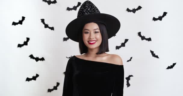 Portré fiatal ázsiai hölgy pózol boszorkány halloween jelmez, mosolygó kamera felett fehér fal papírdenevérek — Stock videók