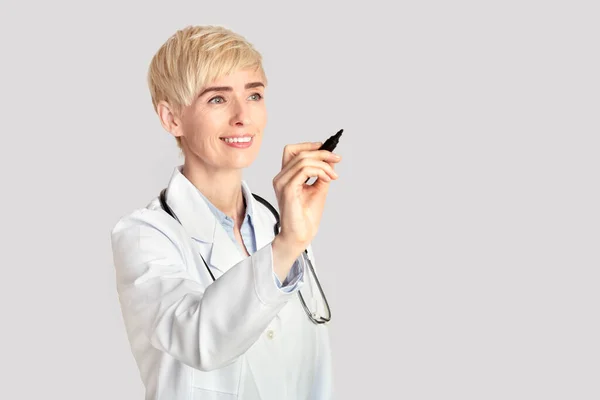 Dorosła lekarka w białym płaszczu ze stetoskopem pisze markerem na wirtualnej tablicy cyfrowej w wolnej przestrzeni — Zdjęcie stockowe