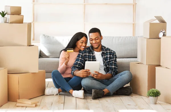 Young black couple using laptop computer and credit card for online shopping at their new home on moving day