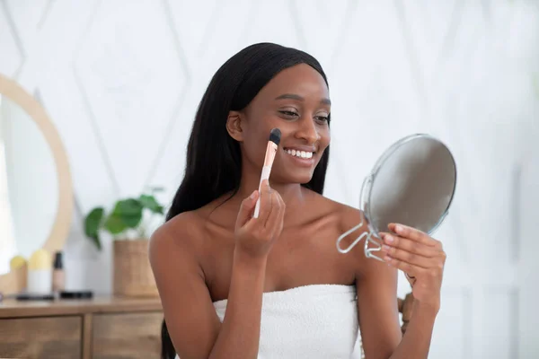 Mujer envuelta en toalla después de la ducha aplicando maquillaje en la cara con cepillo y mirando en el espejo —  Fotos de Stock