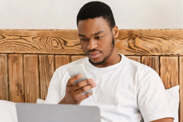 Αφρικανικός άνθρωπος γραπτών μηνυμάτων στο τηλέφωνο χρησιμοποιώντας το φορητό υπολογιστή κάθεται στο κρεβάτι — Φωτογραφία Αρχείου