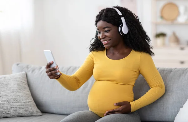 Señora negra embarazada feliz con auriculares usando teléfono inteligente — Foto de Stock