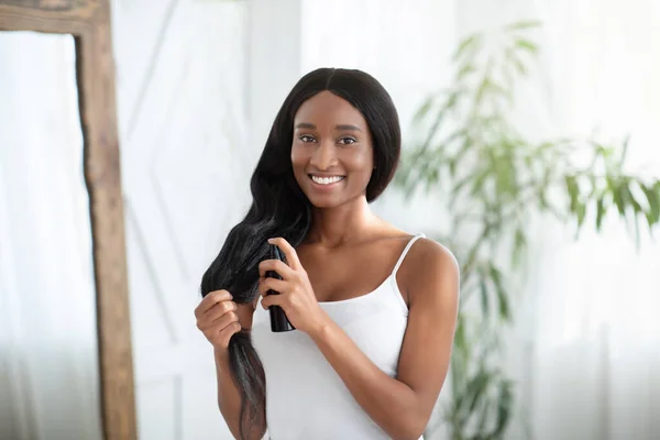 Feliz afro-americana milenar mulher aplicando óleo de cabelo para evitar pontas duplas — Fotografia de Stock