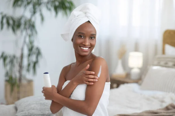 Focus on smiling young african american woman in towel, applies body moisturizer after shower — Stock Photo, Image