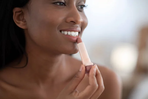 Enfócate en la atractiva dama afroamericana milenaria que aplica lápiz labial desnudo en los labios y se ve en el espejo — Foto de Stock