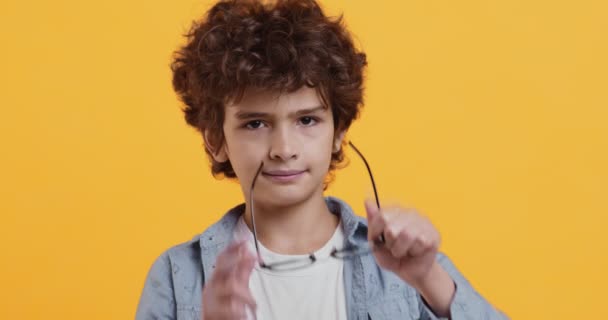 Портрет маленького кудрявого мальчика, надевающего очки и смотрящего в камеру, имеющего проблемы со зрением, оранжевый фон — стоковое видео