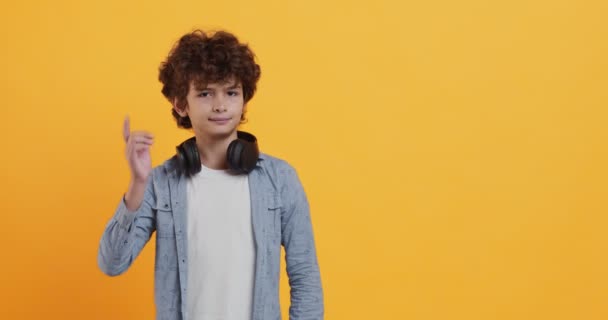 Cool krøllet dreng med hovedtelefoner på halsen indikerer lykkeligt på kamera, flirt koncept – Stock-video