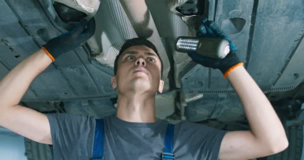 Молодий чоловік у ремонтному сервісі вивчає машину, стоїть під нею зі світлом, крупним планом — стокове відео