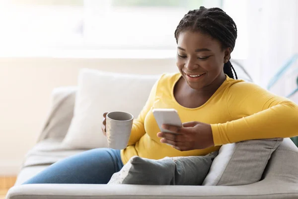 Weekend Leisure. Cheerful black female using smartphone and drinking tea at home — Stock Photo, Image