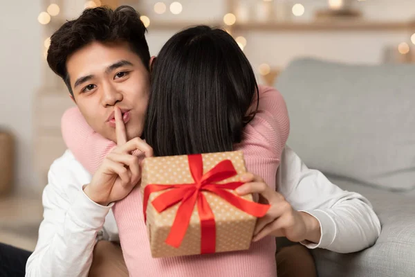 Asian Husband Holding Christmas Gift Behind Wifes Back At Home