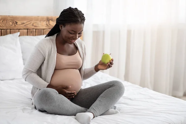 Здоровые закуски. Чернокожая беременная женщина ест зеленое яблоко, отдыхая в постели — стоковое фото