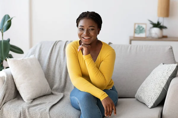 Θετική νεαρή μαύρη γυναίκα που ποζάρει στον καναπέ στο σπίτι, χαμογελώντας στην κάμερα — Φωτογραφία Αρχείου