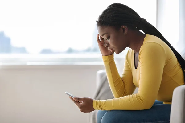 Mujer africana molesta mirando la pantalla del teléfono celular, sentada en el sofá en casa —  Fotos de Stock