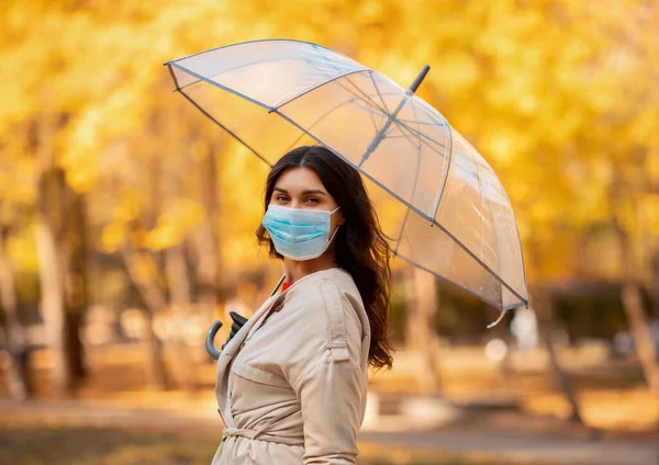 Jolie jeune femme en masque chirurgical sur la promenade sous la pluie au parc le jour de l'automne — Photo