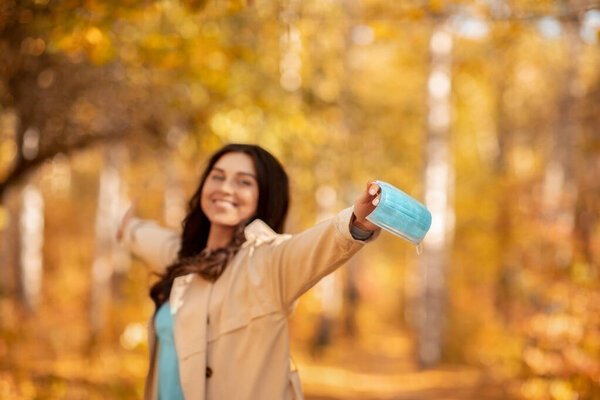 Coronavirus pandemic is over. Happy Caucasian woman taking off surgical mask at fall park, blank space. Selective focus